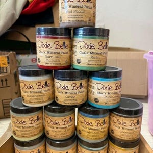 Może przedstawiać: Stos dziesięciu okrągłych plastikowych słoików z farbą kredowo-mineralną Dixie Belle w różnych kolorach, w tym Barn Red, Kudzu, Buttercream, Caviar, Pure Ocean, Farmhouse Green, Spanish Moss, Vintage Duck Egg, Chocolate i Mud Puddle, każdy 237 ml.