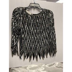 May include: A long-sleeved black top featuring a repeating silver sequin chevron pattern. The garment has a round neckline and a jagged, pointed hemline. A white label with the text St. Honore is visible at the inner back neckline.