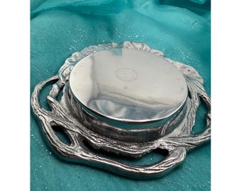 Puede incluir: Un posavasos o base circular de metal plateado con un borde texturizado similar a ramas y un centro liso y reflectante. El objeto metálico tiene una forma orgánica e irregular en el perímetro y una superficie circular plana y pulida.