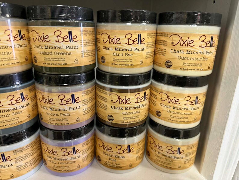 May include: A collection of Dixie Belle Chalk Mineral Paint jars in various colors including Collard Greens, Sand Bar, Cucumber Ice, Secret Path, Chocolate, Buttercream, Lucky Lavender, Clear Coat Flat, and Drop Cloth, each 16 fl oz.