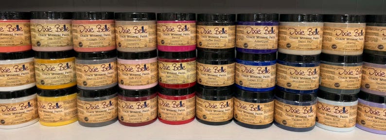 May include: A collection of Dixie Belle Chalk Mineral Paint jars in various colors. The jars are stacked on shelves, showcasing a range of hues from reds and pinks to blues and neutrals. Each jar has a black lid and a label with the brand name.