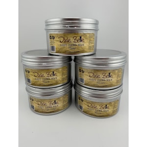 Może przedstawiać: Stos sześciu srebrnych metalowych puszek z woskiem Dixie Belle Best Dang Wax. Każda puszka ma etykietę z nazwą produktu i kolorem: Clear, Black, Brown, Grunge Gray i White. Etykiety są beżowe z czarnym tekstem.