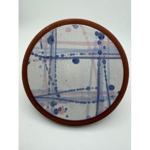 May include: A round ceramic coaster with a brown rim. The center features a white background with a blue and pink splatter design. The coaster is a decorative item for home use.