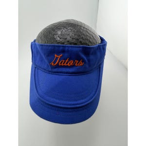 May include: A royal blue visor featuring the word Gators embroidered in orange script across the front panel. The visor has a structured crown and a curved brim, with a black textured dome visible underneath the top portion of the fabric.