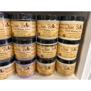 May include: A display of Dixie Belle Chalk Mineral Paint jars in various colors, including Collard Greens, Sand Bar, and Cucumber Ice. Each jar has a black lid and a label with the brand name and paint color.