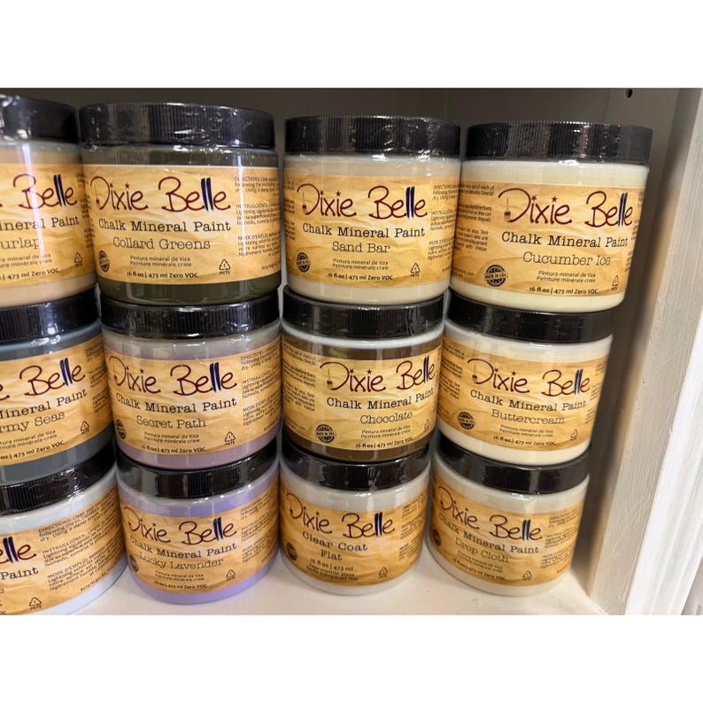 May include: A display of Dixie Belle Chalk Mineral Paint jars in various colors, including Collard Greens, Sand Bar, and Cucumber Ice. Each jar has a black lid and a label with the brand name and paint color.