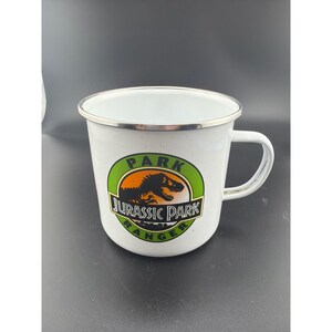 May include: A white enamel mug with a silver rim and a handle. The front features a circular logo with a green border, an orange center, and a black silhouette of a dinosaur. The text reads PARK JURASSIC PARK RANGER in black and white lettering.