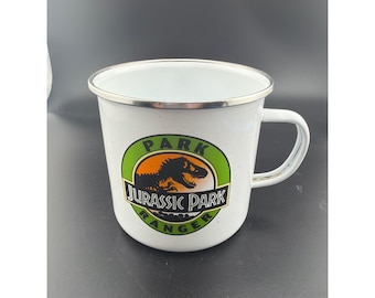 Puede incluir: Una taza de esmalte blanco con borde plateado y asa. El frente presenta un logotipo circular con borde verde, centro naranja y la silueta negra de un dinosaurio. El texto dice PARK JURASSIC PARK RANGER en letras de color negro y blanco.