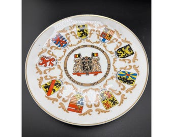 May include: A white porcelain decorative plate with a gold rim, featuring the Belgian coat of arms in the center surrounded by the text SOUVENIR DE BELGIQUE GEDENKENIS VAN BELGIE and nine colorful regional heraldic shields with gold scrollwork.
