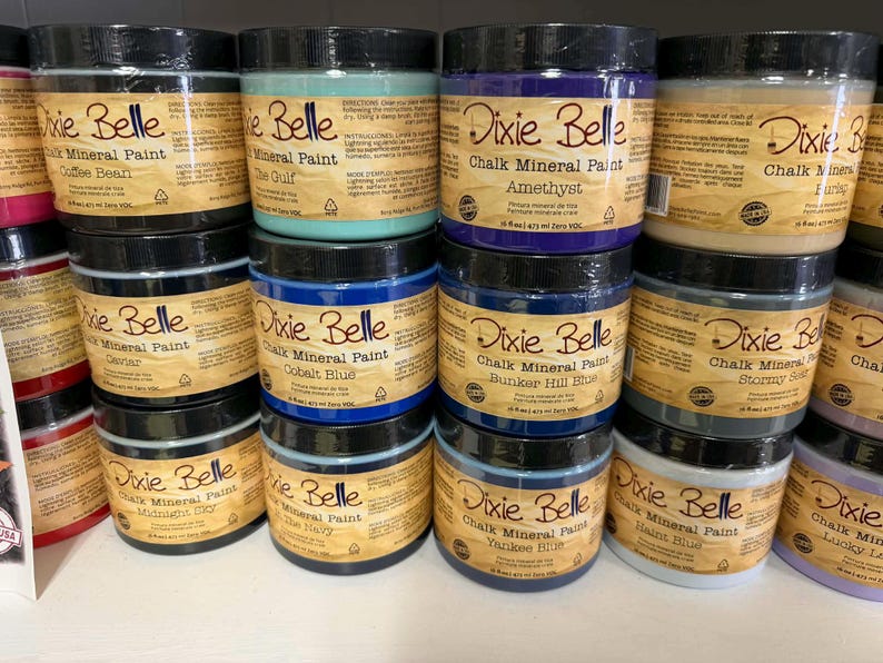 May include: A collection of Dixie Belle Chalk Mineral Paint jars in various colors, including Coffee Bean, Amethyst, and Yankee Blue. Each jar has a black lid and a label with the brand name and paint color. The jars are stacked on shelves.