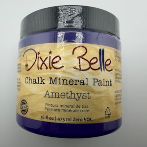 May include: A 16 fl oz jar of Dixie Belle Chalk Mineral Paint in the color Amethyst. The jar features a black lid, a tan label with dark brown text, and the product name clearly printed on the front. The label also indicates Zero VOC.