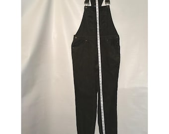 May include: Black corduroy overalls featuring a front bib pocket, adjustable shoulder straps with metal buckles, and side button closures. A white measuring tape is placed vertically over the garment, showing a length of approximately 56 inches.
