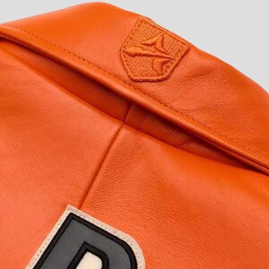 Puede incluir: Primer plano de una chaqueta de cuero naranja. La chaqueta presenta un peque&ntilde;o parche bordado en el hombro y un parche con una letra grande. El cuero tiene una textura suave y un color vibrante.