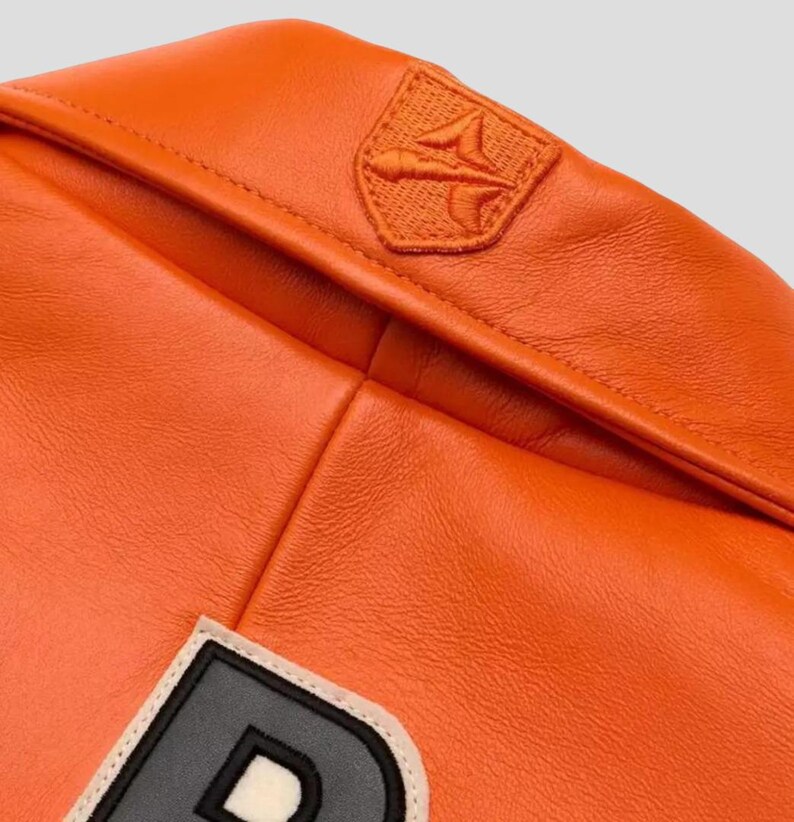 Puede incluir: Primer plano de una chaqueta de cuero naranja. La chaqueta presenta un peque&ntilde;o parche bordado en el hombro y un parche con una letra grande. El cuero tiene una textura suave y un color vibrante.