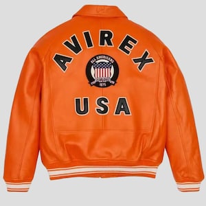 Puede incluir: Chaqueta de cuero naranja brillante con la palabra "AVIREX" arqueada sobre los hombros. La parte trasera presenta un parche circular con las palabras "ALL AMERICANS ESTABLISHED 1975" y un dise&ntilde;o de bandera de EE. UU. La palabra "USA" est&aacute; impresa debajo.