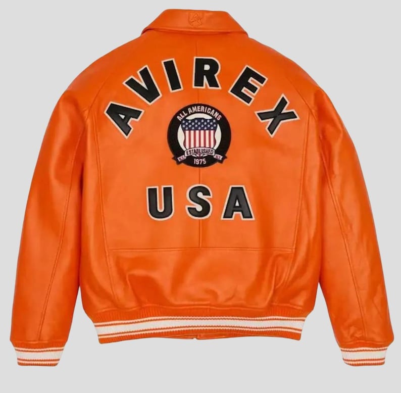 Puede incluir: Chaqueta de cuero naranja brillante con la palabra "AVIREX" arqueada sobre los hombros. La parte trasera presenta un parche circular con las palabras "ALL AMERICANS ESTABLISHED 1975" y un dise&ntilde;o de bandera de EE. UU. La palabra "USA" est&aacute; impresa debajo.