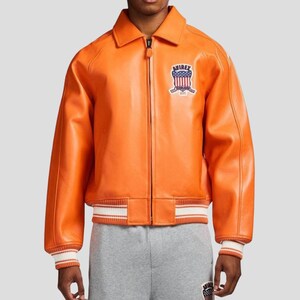 Puede incluir: Chaqueta de cuero naranja con cremallera, cuello y pu&ntilde;os y dobladillo acanalados. La chaqueta presenta un parche en el pecho con la palabra "AVIREX" y un dise&ntilde;o de escudo. Los pu&ntilde;os y el dobladillo tienen rayas blancas.