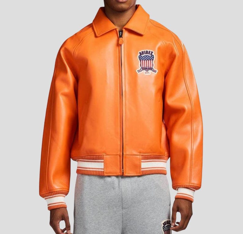 Puede incluir: Chaqueta de cuero naranja con cremallera, cuello y pu&ntilde;os y dobladillo acanalados. La chaqueta presenta un parche en el pecho con la palabra "AVIREX" y un dise&ntilde;o de escudo. Los pu&ntilde;os y el dobladillo tienen rayas blancas.