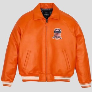Puede incluir: Chaqueta de cuero naranja brillante con cremallera, cuello y parche en el pecho. La chaqueta tiene dos bolsillos delanteros y una banda a rayas blancas y naranjas en la cintura y los pu&ntilde;os. El parche presenta un dise&ntilde;o con elementos rojos, blancos y azules.