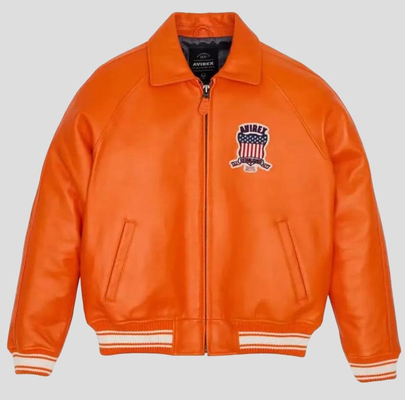 Puede incluir: Chaqueta de cuero naranja brillante con cremallera, cuello y parche en el pecho. La chaqueta tiene dos bolsillos delanteros y una banda a rayas blancas y naranjas en la cintura y los pu&ntilde;os. El parche presenta un dise&ntilde;o con elementos rojos, blancos y azules.