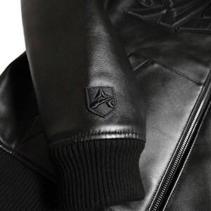 May include: Close-up of a black leather jacket. The jacket features a ribbed cuff, a small emblem on the sleeve, and a zipper. The leather has a slight sheen. The jacket appears to be new.