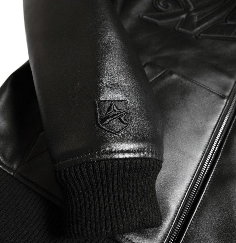 May include: Close-up of a black leather jacket. The jacket features a ribbed cuff, a small emblem on the sleeve, and a zipper. The leather has a slight sheen. The jacket appears to be new.