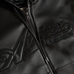 May include: Close-up of a black leather jacket with a zipper closure. The jacket features the embroidered text "Avirex USA" in a bold, stylized font. The leather appears smooth and the stitching is detailed.