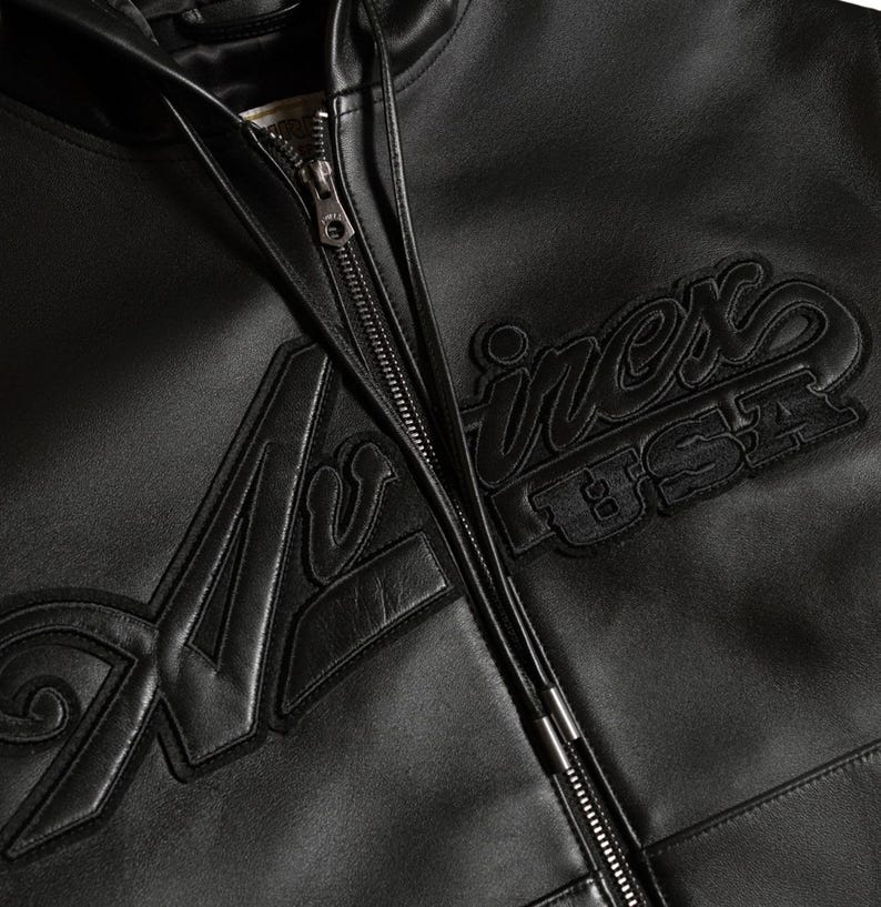 May include: Close-up of a black leather jacket with a zipper closure. The jacket features the embroidered text "Avirex USA" in a bold, stylized font. The leather appears smooth and the stitching is detailed.