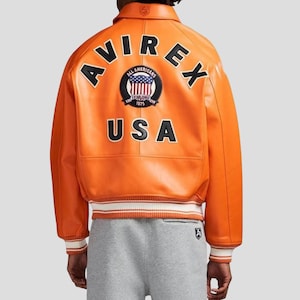 Puede incluir: Chaqueta de cuero naranja con la palabra "AVIREX" en letras negras en la espalda. Debajo, un parche circular con las palabras "ALL AMERICANS" y "ESTABLISHED 1975" que rodea un dise&ntilde;o de la bandera estadounidense. La palabra "USA" est&aacute; debajo.