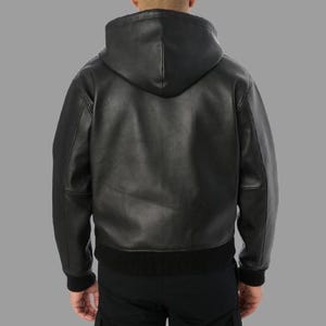 May include: Black leather hooded jacket with a ribbed hem and cuffs. The jacket has a full-length zipper and a hood. The model is wearing black pants. The jacket is made of a smooth, durable leather material.