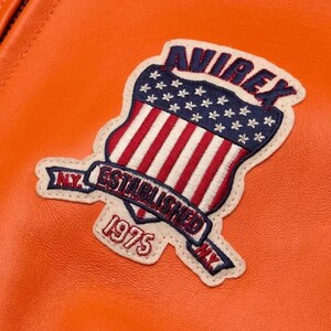 Puede incluir: Primer plano de una chaqueta de cuero naranja con un parche con el texto "AVIREX" sobre un dise&ntilde;o de escudo con rayas rojas y blancas y estrellas azules. El parche tambi&eacute;n incluye el texto "ESTABLISHED 1975 NY".