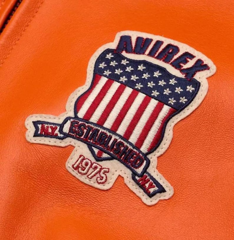 Puede incluir: Primer plano de una chaqueta de cuero naranja con un parche con el texto "AVIREX" sobre un dise&ntilde;o de escudo con rayas rojas y blancas y estrellas azules. El parche tambi&eacute;n incluye el texto "ESTABLISHED 1975 NY".