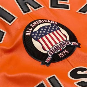 Puede incluir: Primer plano de una chaqueta de cuero naranja con letras negras y blancas. Un parche circular presenta un dise&ntilde;o de escudo con la bandera estadounidense y las palabras "ALL AMERICANS ESTABLISHED 1975 NY."