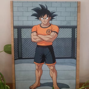 Puede incluir: Ilustración enmarcada de un personaje musculoso de la serie de anime Dragon Ball. El personaje tiene el pelo negro puntiagudo, lleva una camiseta naranja con un símbolo japonés y pantalones cortos azul oscuro con ribetes naranjas. La obra está sobre un fondo gris.