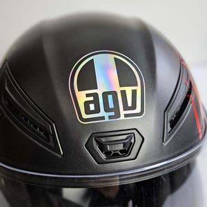 Può includere: Casco da moto nero opaco con il logo "AGV" in un quadrato con una striscia arcobaleno. Il casco ha una visiera trasparente e prese d'aria. Dettagli rossi sono visibili sul lato.