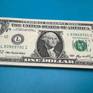 Pode incluir: Uma pilha de notas de um dólar americano. Cada nota apresenta um retrato de George Washington e o texto "ONE DOLLAR". As notas são verdes e pretas, com o texto "THE UNITED STATES OF AMERICA" no topo.