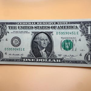 Pode incluir: Uma nota de um dólar dos Estados Unidos, com um retrato de George Washington. A nota exibe o texto "The United States of America" e "One Dollar". O número de série é visível.