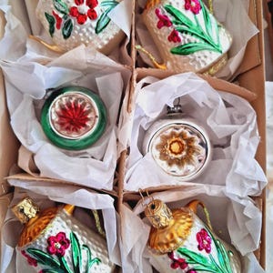 May include: A set of vintage Christmas ornaments in a cardboard box. The ornaments include teacup-shaped and round glass baubles. The teacups are white with gold accents and painted red flowers and green leaves. The round ornaments have red and gold designs.