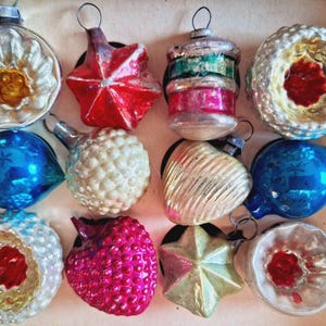 May include: A collection of vintage Christmas ornaments in various shapes and colors. The ornaments include a red star, a blue teardrop, and a white textured ball. Other ornaments are silver, red, and green. The ornaments are in a cardboard box.