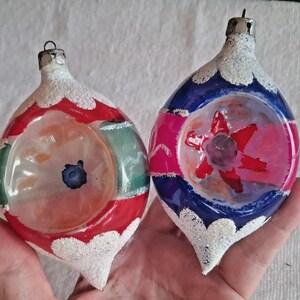 May include: Two vintage Christmas ornaments. One is red, green, and white, the other is blue, pink, and white. Both have a teardrop shape with a silver cap and a white, glittery base. The ornaments have a clear center with painted designs.