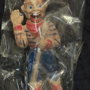 May include: A vintage ventriloquist dummy toy, packaged in clear plastic. The doll has a painted face with blue eyes, a red scarf, and blue pants. Its arms and legs are articulated, and it is wearing red and blue boots.