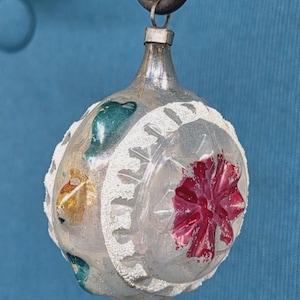 May include: Vintage Christmas ornament with a silver finish, featuring a red flower and accents of gold and teal. The ornament has a white, textured border and is suspended from a metal loop. A classic holiday decoration.