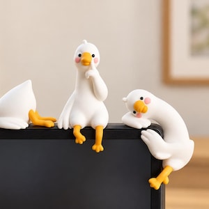 May include: Three white goose figurines with orange beaks and feet and pink cheeks. One goose lies flat, one sits upright with a wing to its beak, and one hangs over the edge. These decorative figures are positioned along the top of a black monitor.