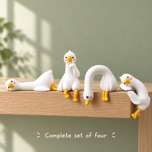 Op de afbeelding: Een complete set van vier witte ganzenbeeldjes met gele snavels en voeten, voorzien van roze wangen. Elk beeldje heeft een andere houding: één ligt plat, één zit met een vinger bij de snavel, één buigt voorover en één rust op de zijkant.