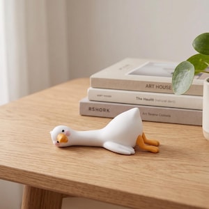 May include: A white goose figurine with an orange beak and orange feet, depicted in a prone position with its head resting on the surface. The goose has small black eyes and pink circular markings on its cheeks, appearing as a smooth, solid object.