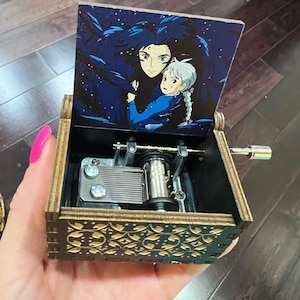 May include: A wooden music box with an anime illustration on the lid, depicting two characters against a starry night sky. The box features a decorative pattern and a silver-coloured crank. The mechanism is visible.