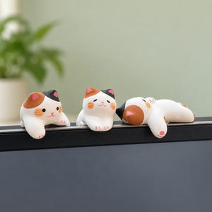 May include: Three small calico cat figurines with white, orange, and black markings. Each cat has a different pose, with one looking forward, one sitting upright, and one lying down, all designed to perch on the edge of a flat surface.