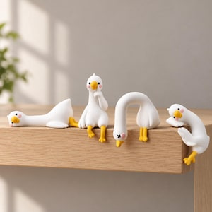 Könnte beinhalten: Vier kleine weiße Gänsefiguren mit gelben Schnäbeln und Füßen, jede in einer anderen Pose. Eine liegt flach, eine sitzt aufrecht, eine beugt sich über die Kante und eine hängt an der Seite. Sie haben rosa Wangendetails.