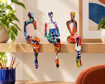 Op de afbeelding: Drie abstracte mensvormige sculpturen zittend op een richel, elk voorzien van een levendig, meerkleurig graffiti-patroon met gedurfde spatten oranje, blauwe, rode en zwarte verf over hun gladde, gestileerde vormen.