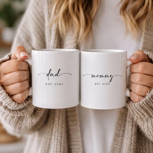 May include: Two white ceramic mugs featuring black printed text. The left mug displays the word dad in a cursive script above the text EST. 2026. The right mug displays the word mommy in a cursive script above the text EST. 2026.